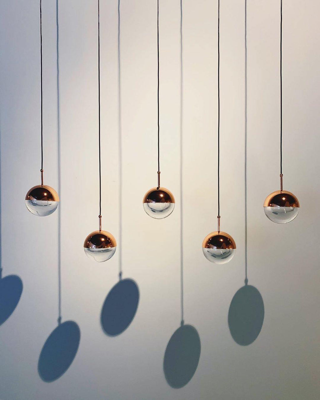 Five rose gold Clérachic globe pendants hung at varied heights against a pale wall, casting round blue shadows.