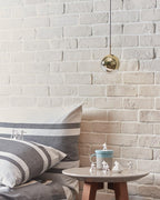 Single gold-finish Clérachic globe pendant against a white brick wall, above a round side table with a mug and figurines.