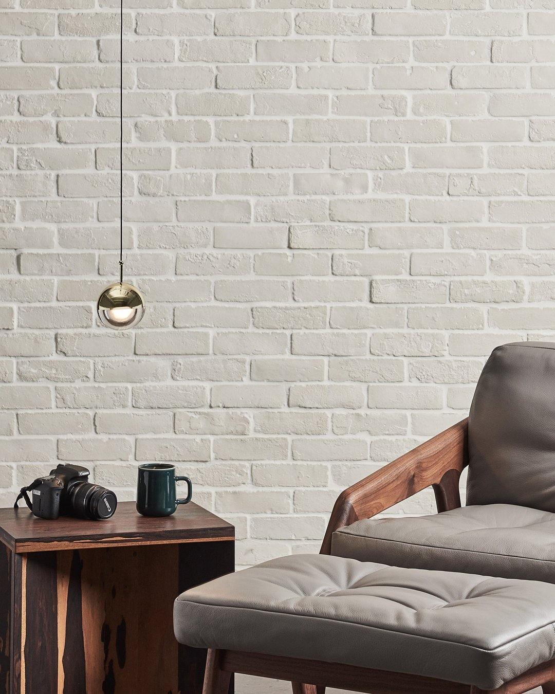 Single gold-finish Clérachic globe pendant against white brick, above a leather armchair, a wooden table and a camera.