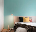Single rose gold Clérachic globe pendant on a turquoise wall, above a bed with a dark throw and yellow cushion.