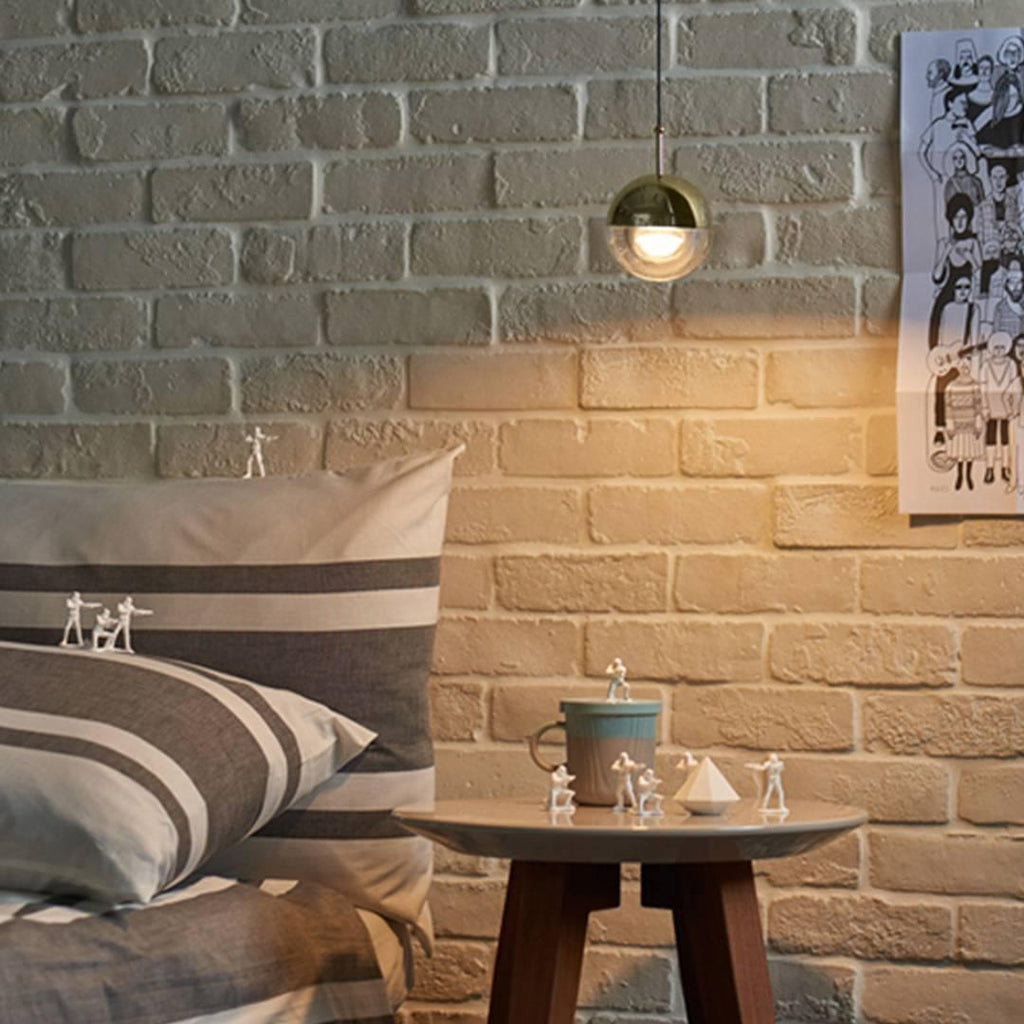 A gold-finish Clérachic globe pendant glowing against an exposed brick wall, with a striped cushion and a round side table below.