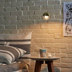 A gold-finish Clérachic globe pendant glowing against an exposed brick wall, with a striped cushion and a round side table below.