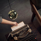 Gold mirror-finish Clérachic globe pendant above a hand feeding paper into a vintage typewriter on a dark desk.