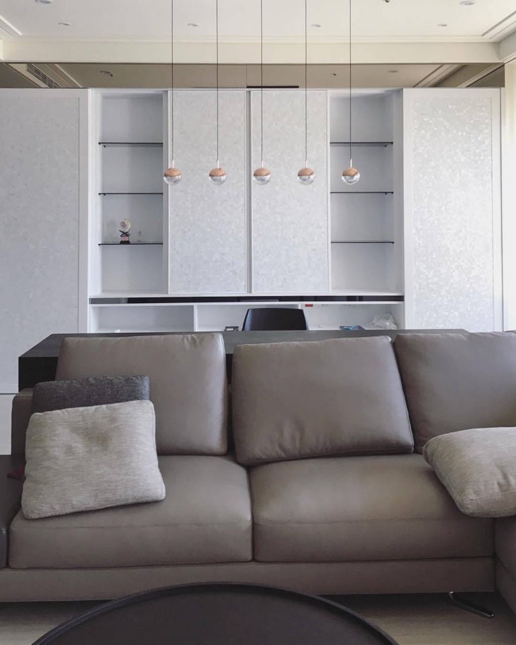 A row of five small rose gold Clérachic globe pendants hung over a grey sofa, in front of white cabinets and open shelves.