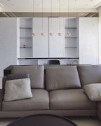A row of five small rose gold Clérachic globe pendants hung over a grey sofa, in front of white cabinets and open shelves.
