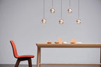 Five rose gold Clérachic globe pendants at staggered heights above a pale wooden table and an orange chair.