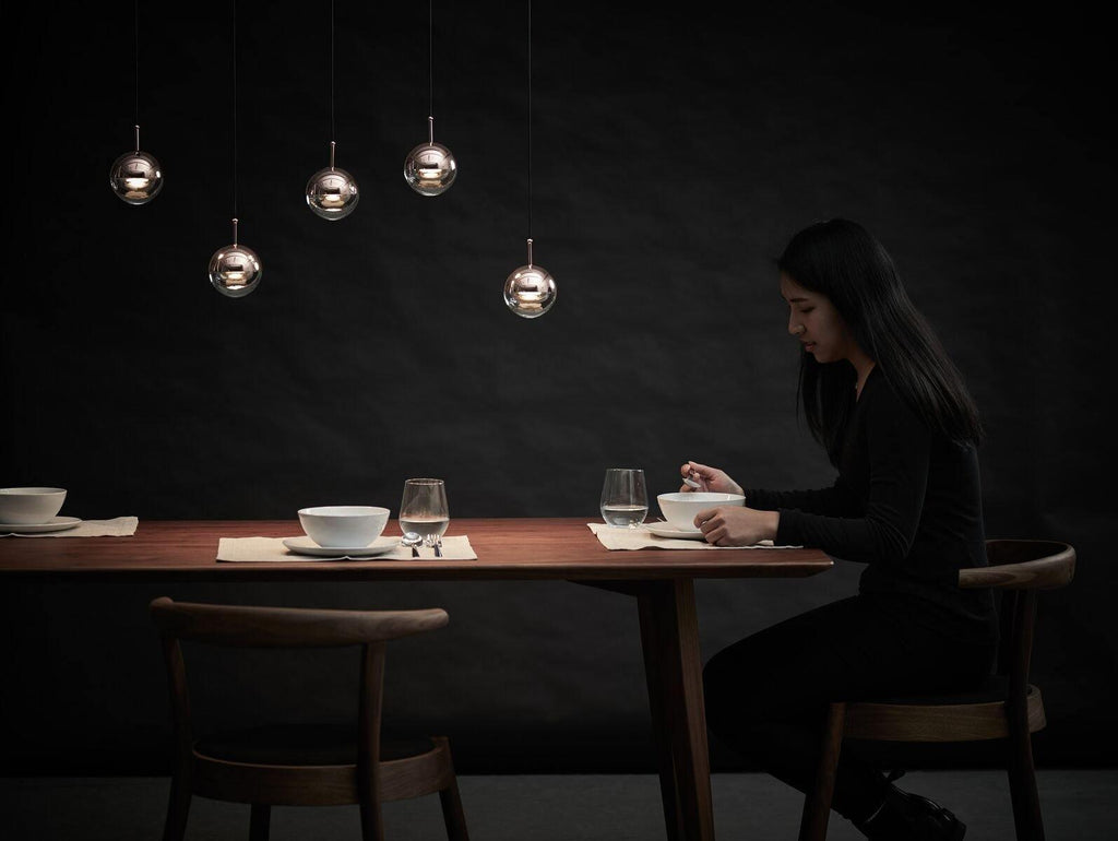 Five small Clérachic globe pendants against a dark wall, over a wooden table where a woman sits eating from a bowl.