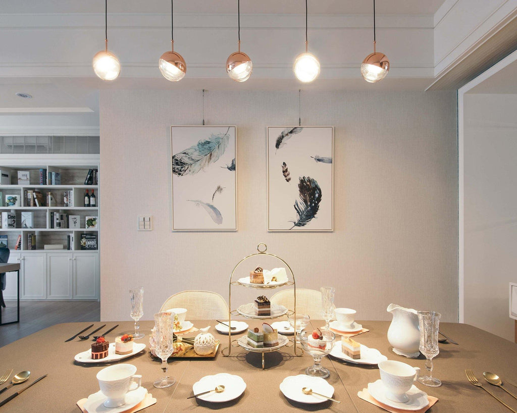 The same row of five Clérachic globe pendants lit warmly, with a warmer glow over the afternoon-tea table below.