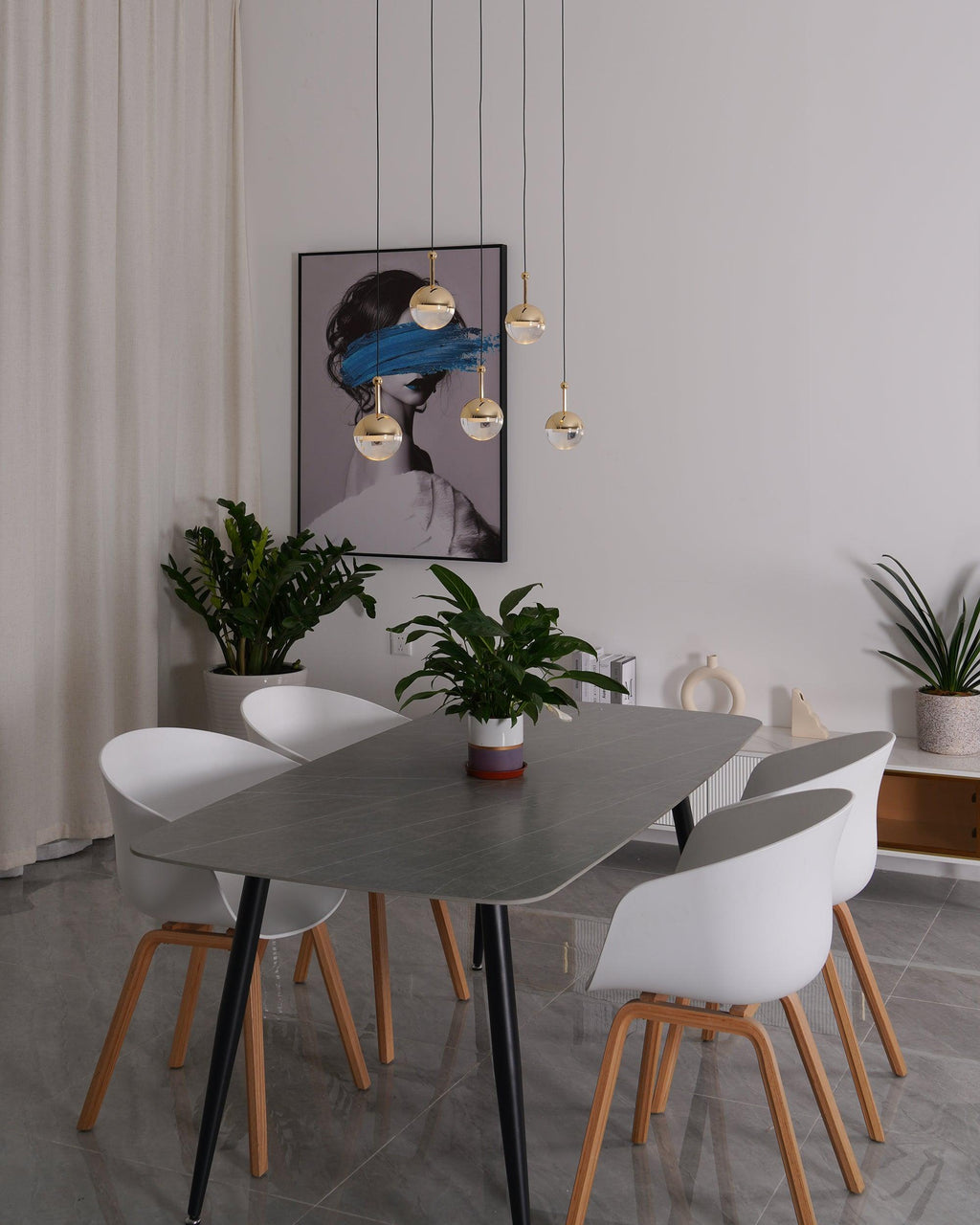 Unlit gold-finish Clérachic globe pendants hung at varied heights in front of a framed portrait, above a grey dining table.