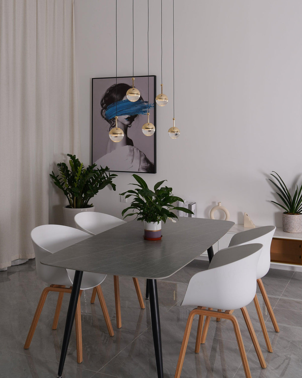 Unlit gold-finish Clérachic globe pendants hung at varied heights in front of a framed portrait, above a grey dining table.