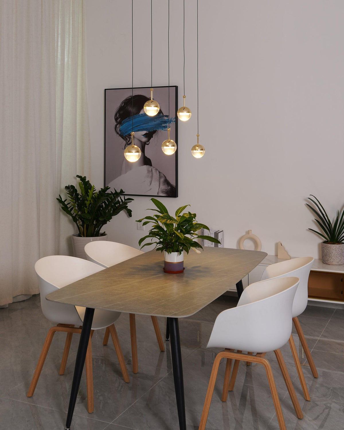 Lit gold-finish Clérachic globe pendants in front of a framed portrait, above a grey table with white chairs and a plant.