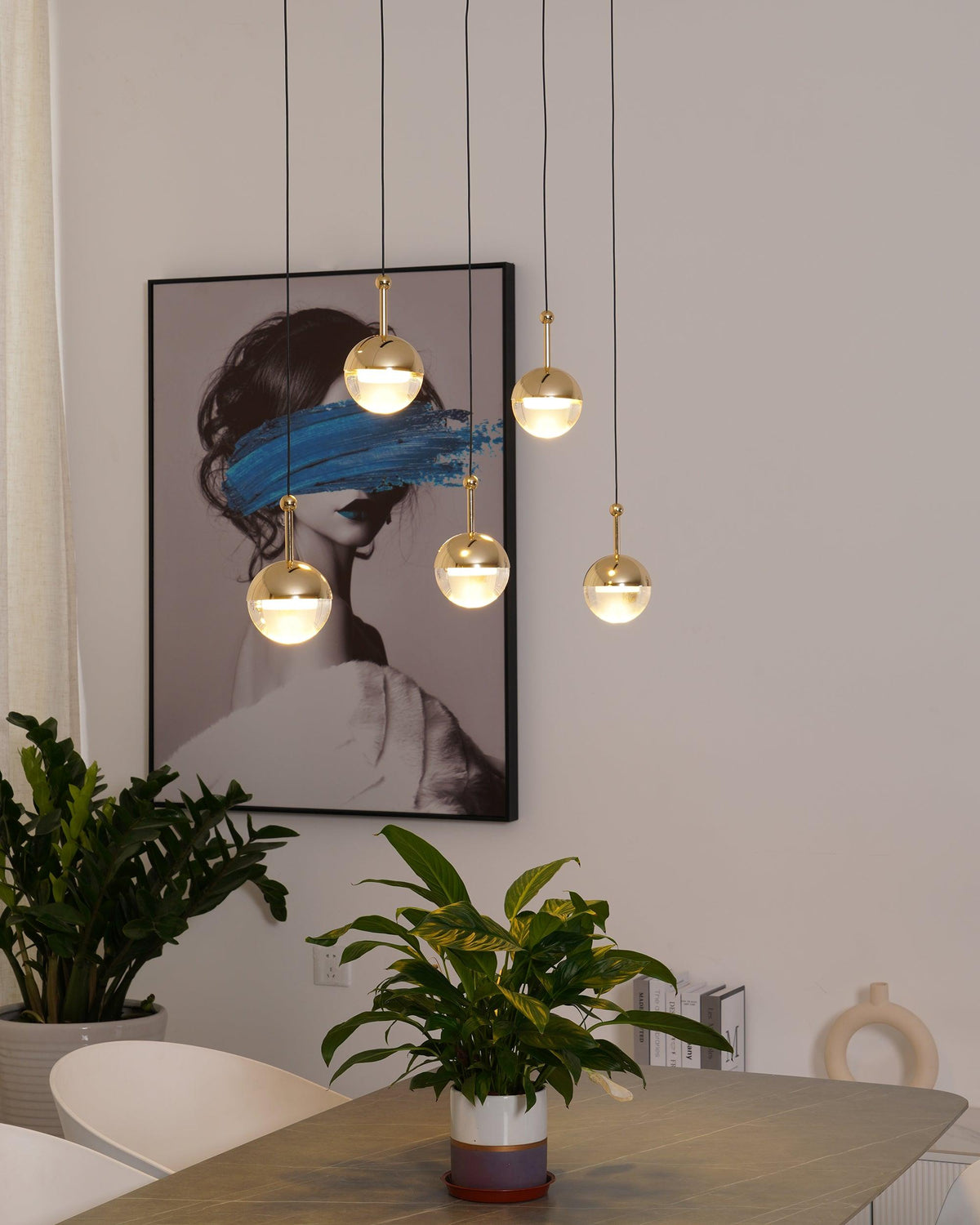 Close view of lit gold-finish Clérachic globe pendants beside a framed portrait with a blue brushstroke, above a potted plant.