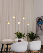 Five lit gold-finish Clérachic globe pendants at staggered heights in front of a cream curtain, above a dark dining table.