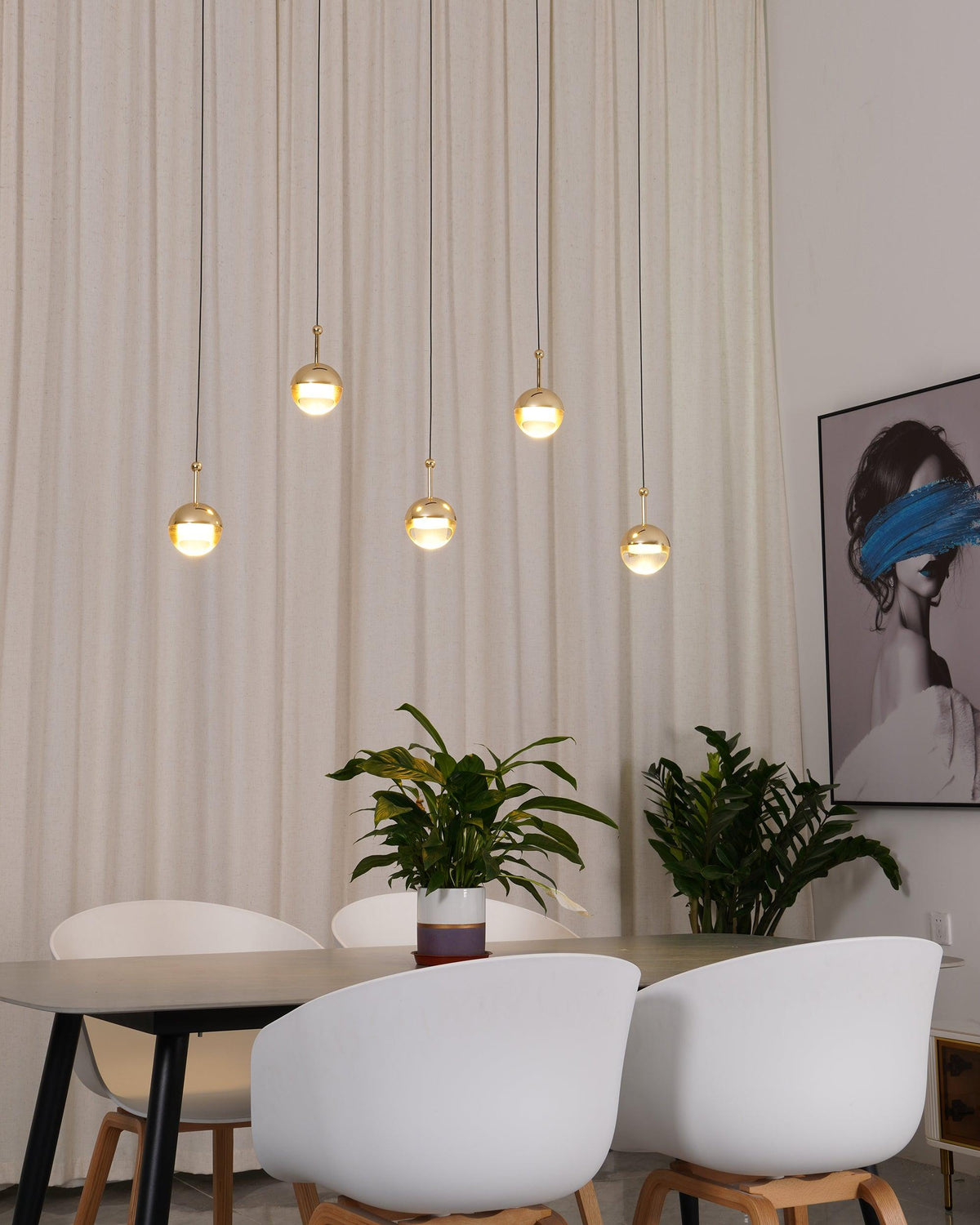 Five lit gold-finish Clérachic globe pendants at staggered heights in front of a cream curtain, above a dark dining table.