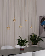 Five unlit gold-finish Clérachic globe pendants at different heights against a cream curtain, above a table and a leafy plant.