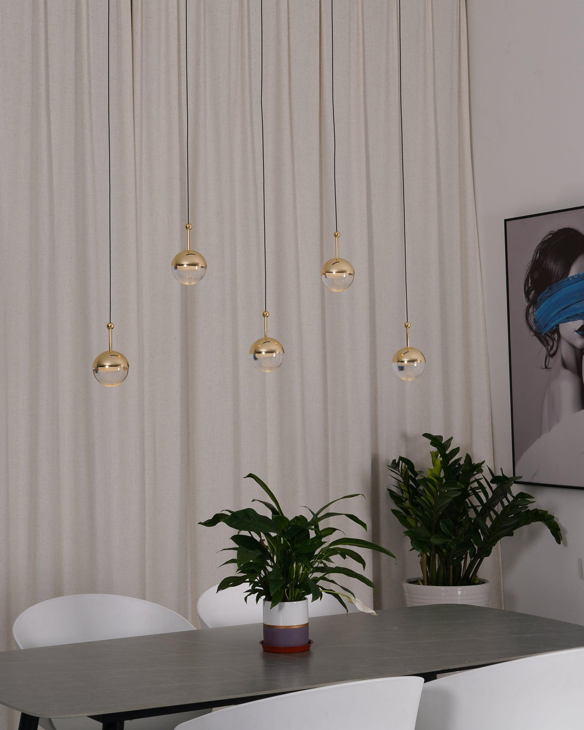 Five unlit gold-finish Clérachic globe pendants at different heights against a cream curtain, above a table and a leafy plant.