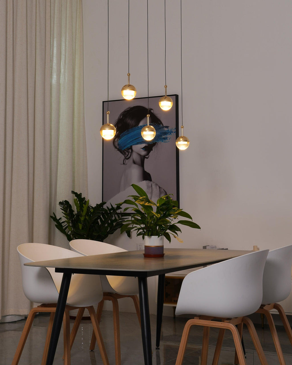 Lit gold-finish Clérachic globe pendants at varied heights by a framed portrait, above a dark table with white chairs.