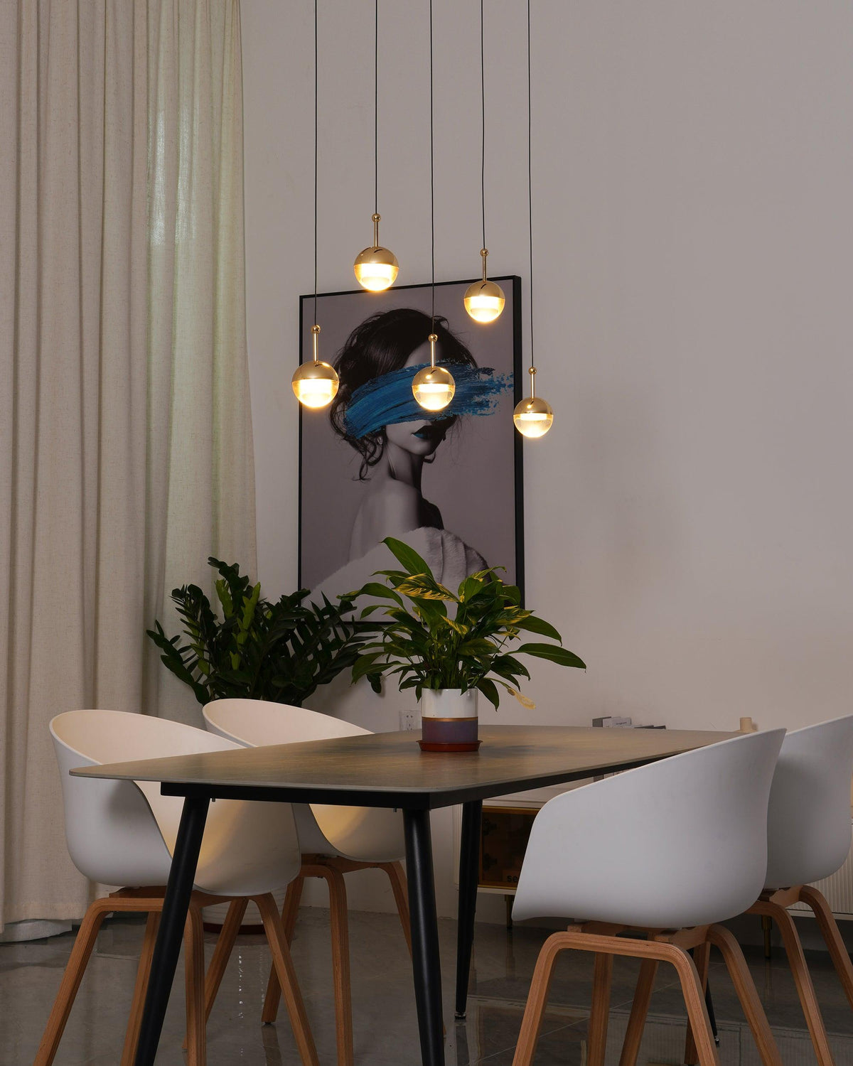 Lit gold-finish Clérachic globe pendants at varied heights by a framed portrait, above a dark table with white chairs.