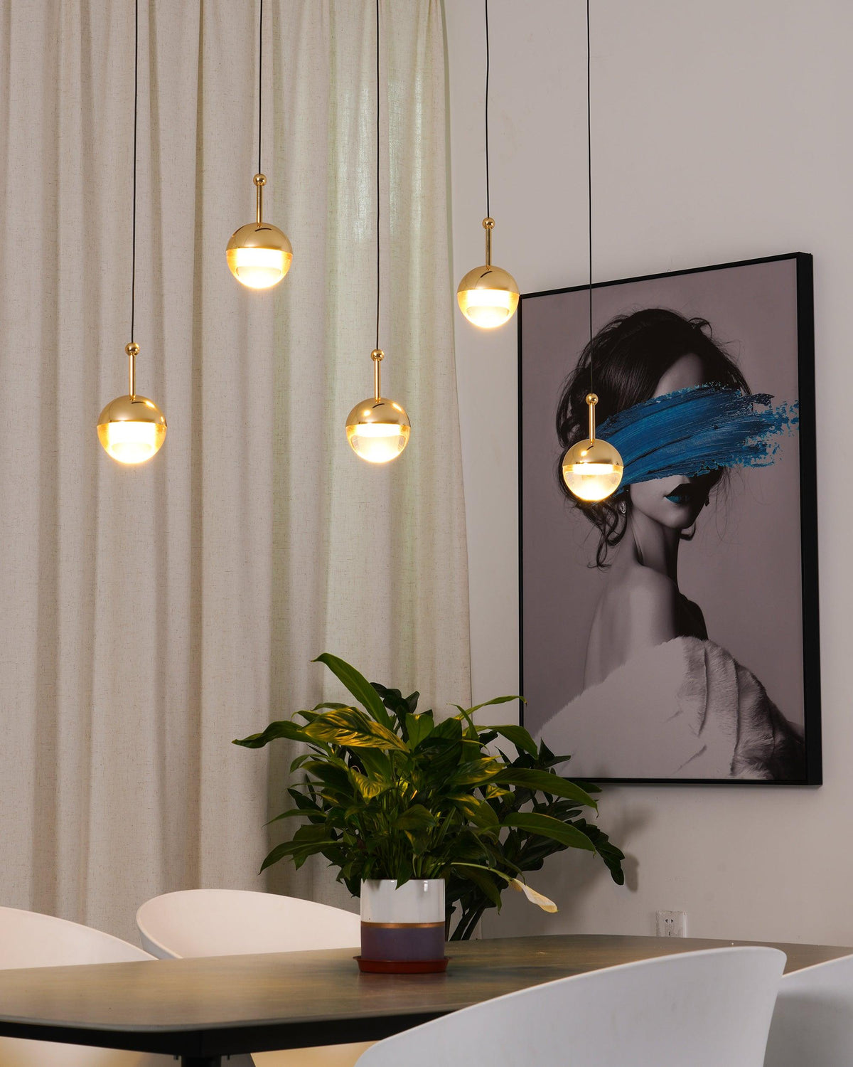 Lit gold-finish Clérachic globe pendants at varied heights in front of a cream curtain and a framed portrait, above a dark table.