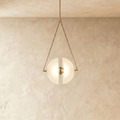 Clérachic pendant with two frosted glass half-discs glowing warm inside a slim triangular brass frame, against limewash.