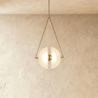 Clérachic pendant with two frosted glass half-discs glowing warm inside a slim triangular brass frame, against limewash.