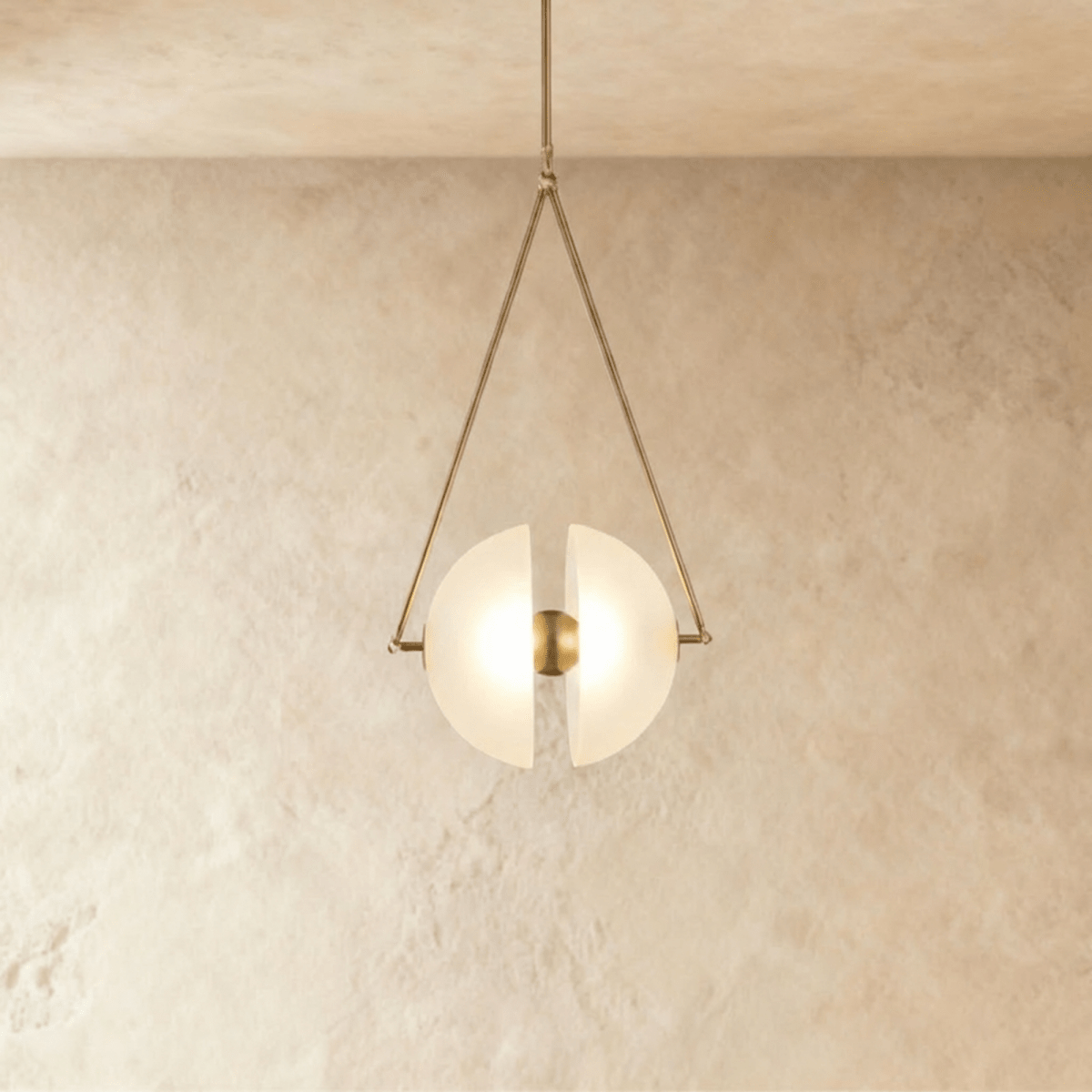 Clérachic pendant with two frosted glass half-discs glowing warm inside a slim triangular brass frame, against limewash.