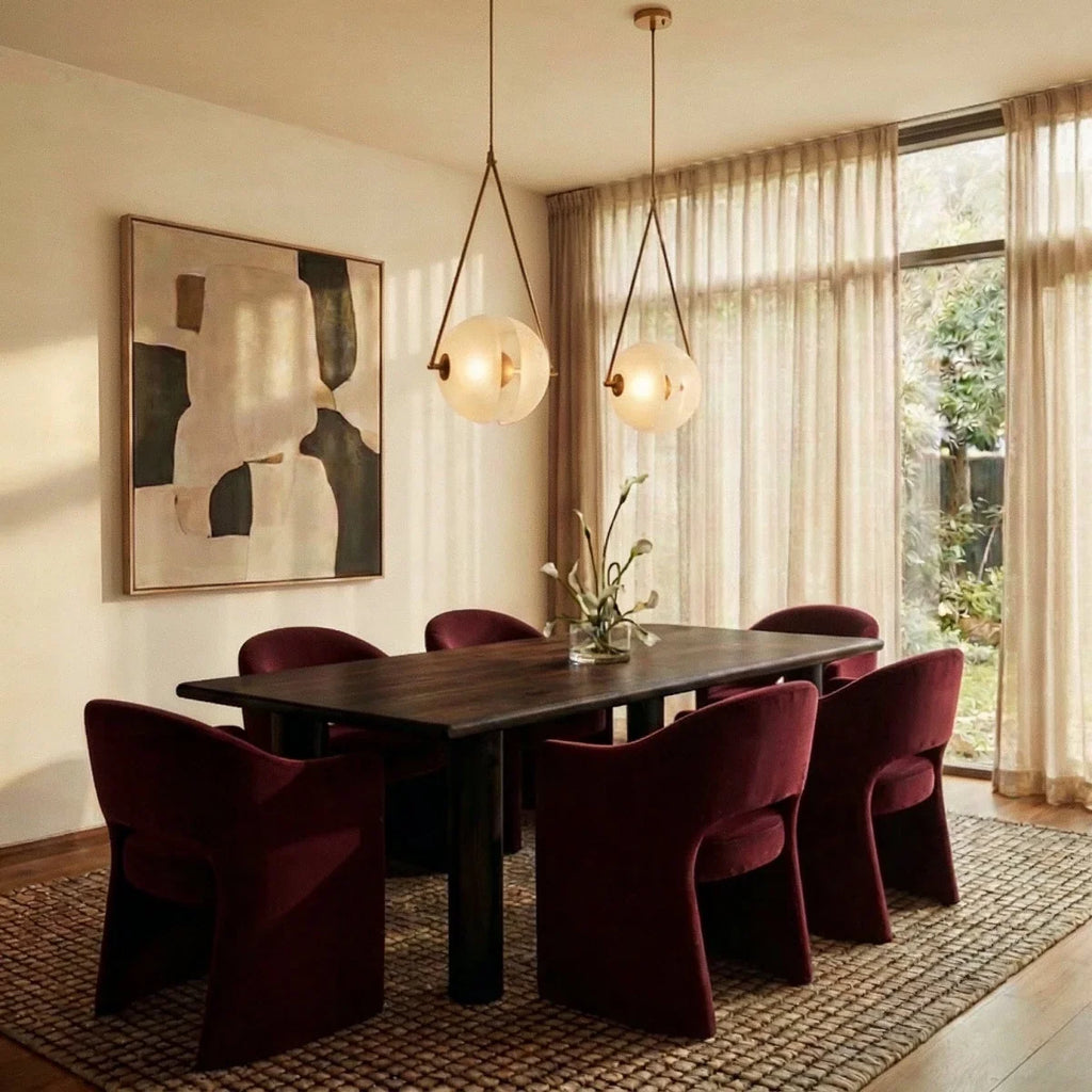 Clérachic two brass-framed frosted glass pendants lit warm above a dark dining table with burgundy chairs and sheer curtains.
