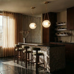 Clérachic two brass-framed frosted glass pendants lit warm above a stone-clad kitchen island with upholstered stools.