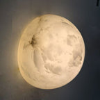 A Clérachic Half-Dome Alabaster Wall Sconce lit on a grey wall at night, heavy cloud and crack figuring visible through the stone.