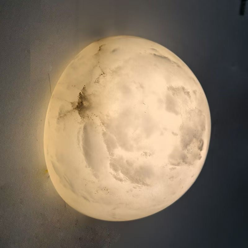 A Clérachic Half-Dome Alabaster Wall Sconce lit on a grey wall at night, heavy cloud and crack figuring visible through the stone.