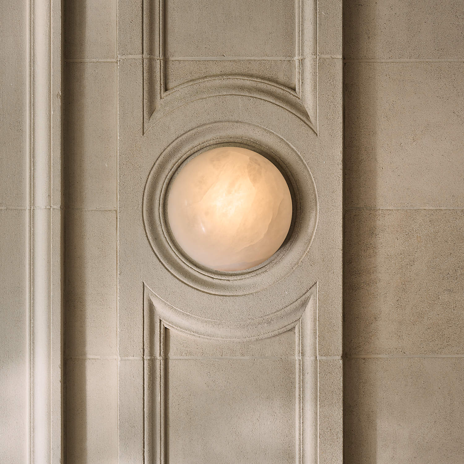 Clérachic Half-Dome Alabaster Wall Sconce lit inside a carved limestone panel, the warm stone face seated in a circular recess.