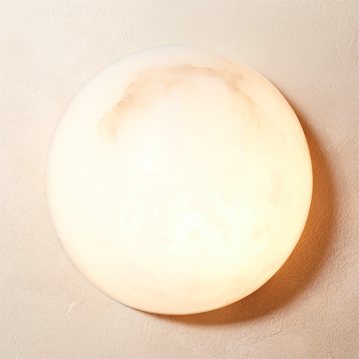 The Clérachic Half-Dome Alabaster Wall Sconce glowing warm on a plaster wall, caramel veining banding the top of the lit dome.