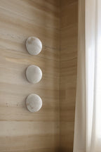 Three Clérachic Half-Dome Alabaster Wall Sconces stacked down a travertine wall beside a sheer curtain in daylight.