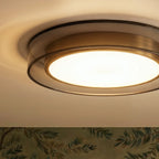 Close view of the Clérachic halo ring ceiling light's brass trim, glowing above leaf-patterned wallpaper edge.