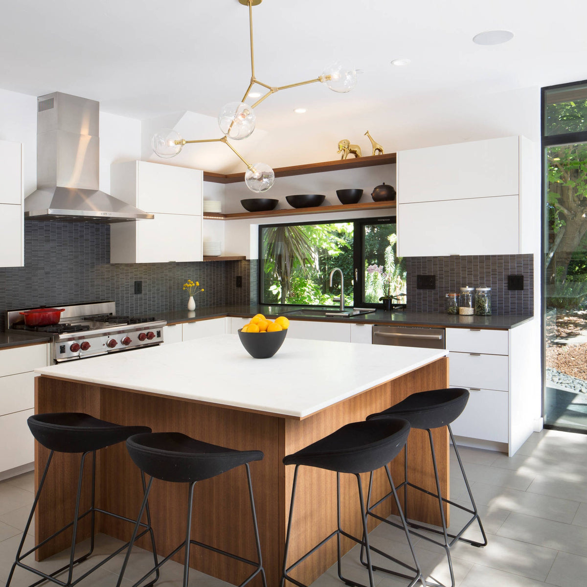 A gold Clérachic chandelier with clear globes hangs above a white kitchen island, with black stools and a window behind.