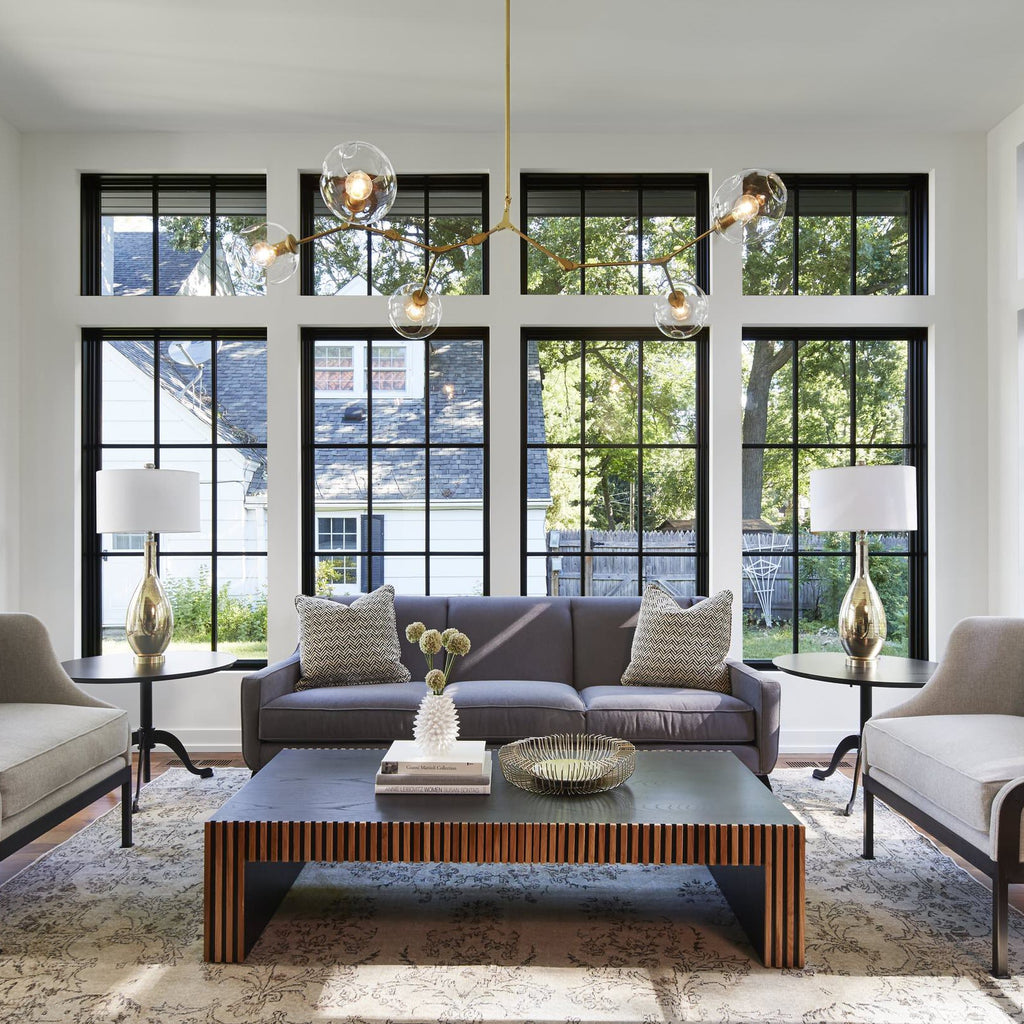 A Clérachic chandelier with clear globes and gold arms hangs in front of a wall of black-framed windows above a grey sofa.