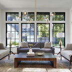 A Clérachic chandelier with clear globes and gold arms hangs in front of a wall of black-framed windows above a grey sofa.