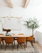 A Clérachic chandelier with gold arms hangs between pale wooden beams over a dining table with tan chairs and an olive tree.