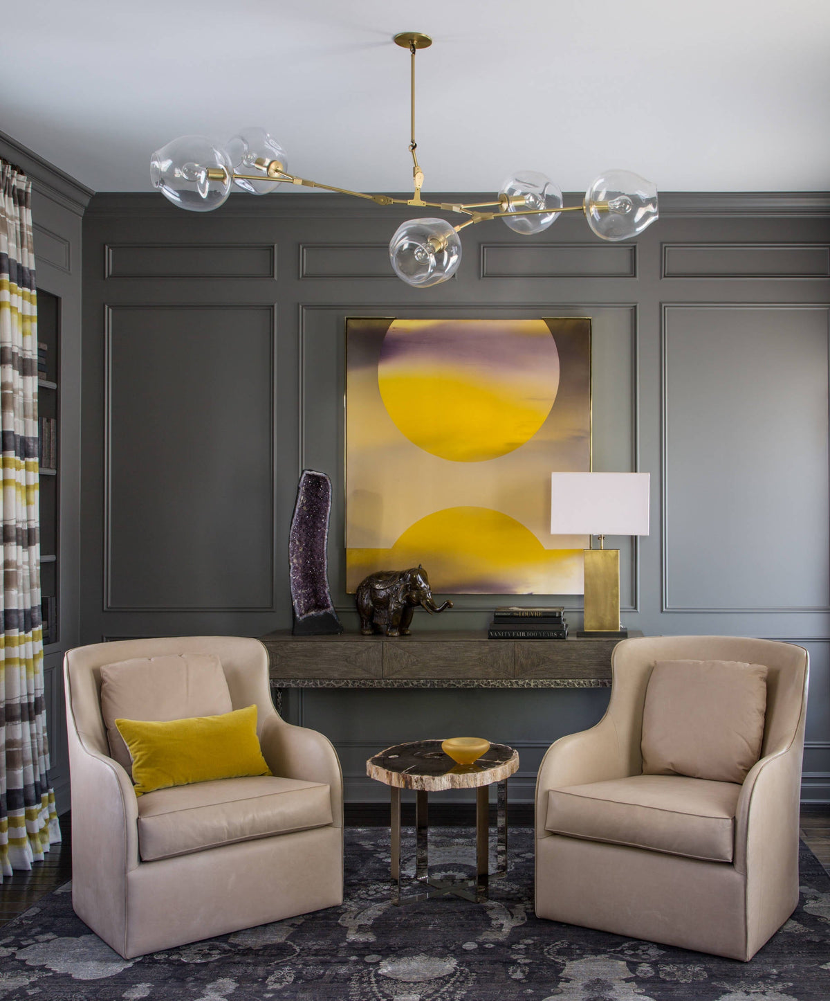 Clear globes along a gold arm form this Clérachic chandelier, hanging in a grey panelled room with two cream armchairs below.