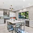 A small gold Clérachic chandelier hangs above a marble kitchen island with metal-framed stools, with a grey tiled wall behind.