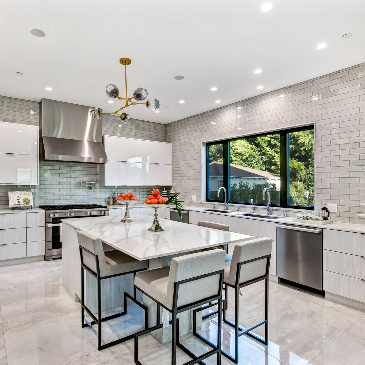 A small gold Clérachic chandelier hangs above a marble kitchen island with metal-framed stools, with a grey tiled wall behind.
