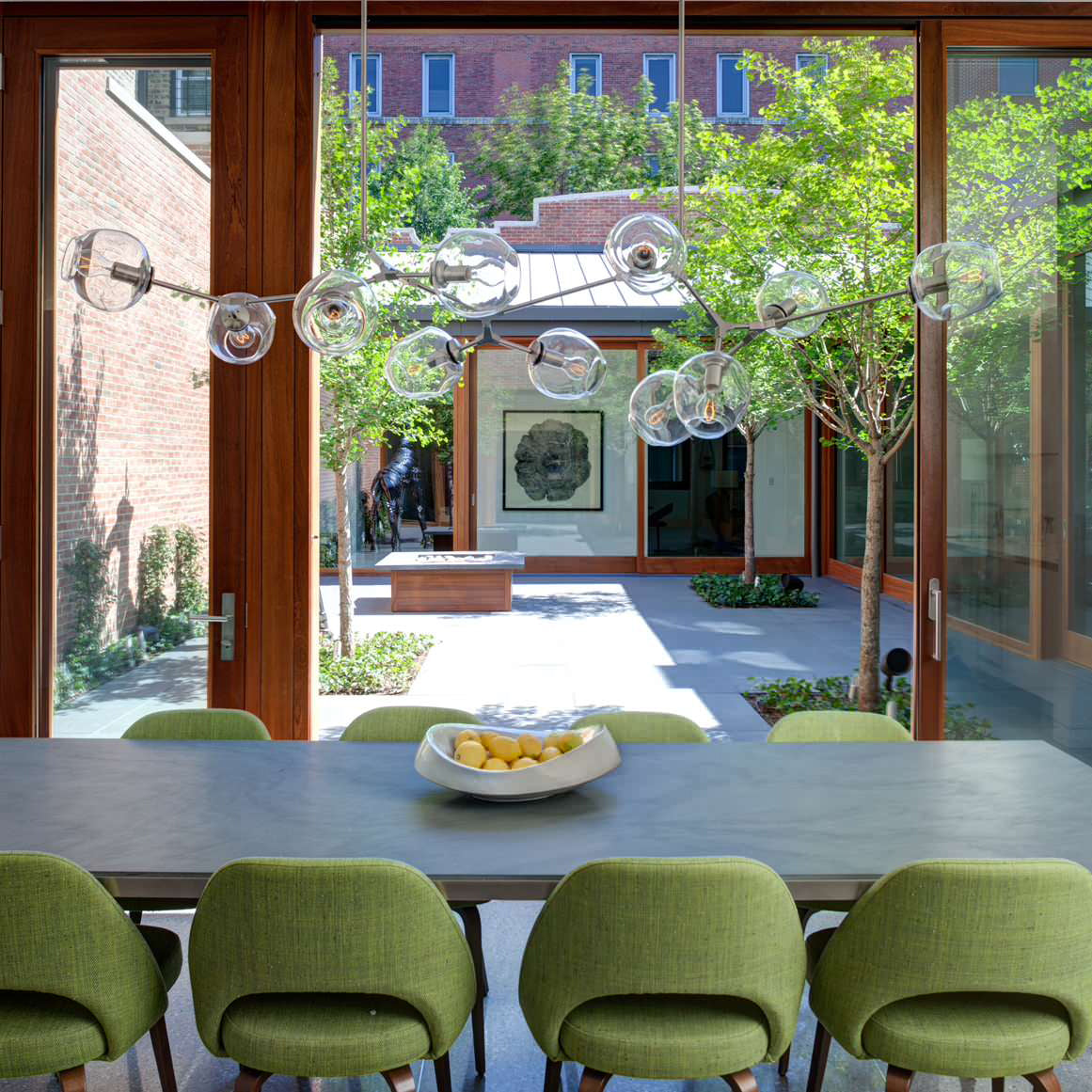 A long run of clear glass globes from this Clérachic chandelier hangs before open doors to a courtyard, above green dining chairs.