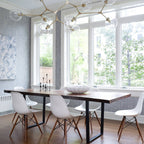 Clear globes along pale metal arms of this Clérachic chandelier hang before tall white windows above a long wooden table.