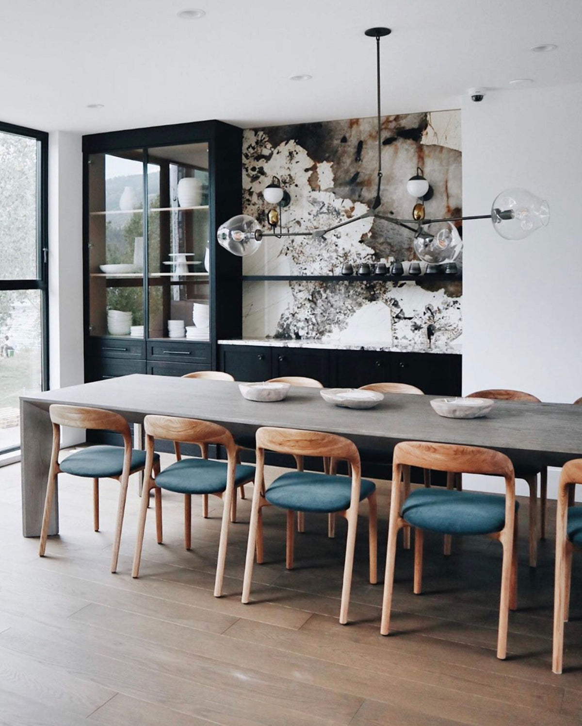 This Clérachic chandelier with black arms and clear globes hangs over a long grey table, with teal-cushioned wooden chairs below.