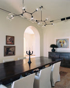 A black-framed Clérachic chandelier with clear globes spreads across the ceiling above a dark dining table and white chairs.