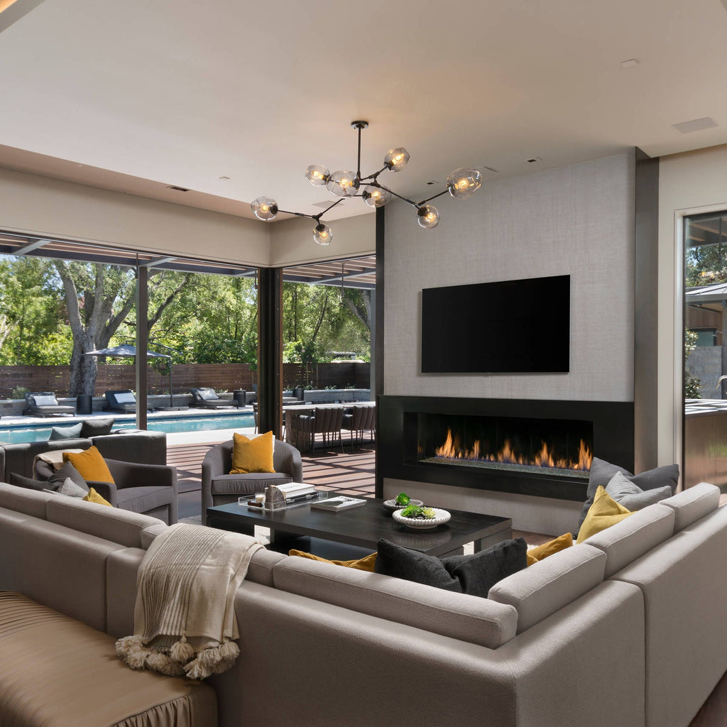 A Clérachic chandelier with black arms and clear globes hangs over a large sectional sofa and a linear fireplace in a wide living room.