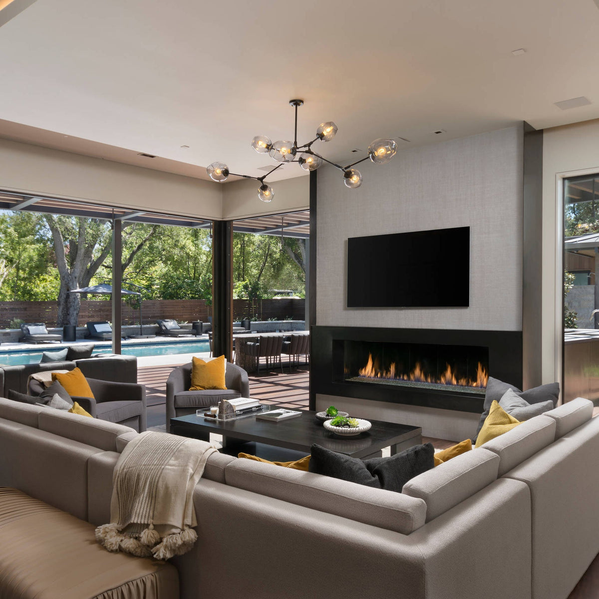 A Clérachic chandelier with black arms and clear globes hangs over a large sectional sofa and a linear fireplace in a wide living room.