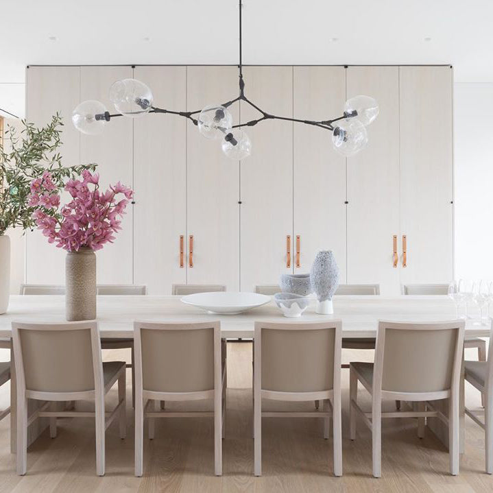 A long Clérachic chandelier with black arms and clear globes spans a white kitchen wall, over chairs with pale grey cushions.