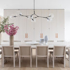 A long Clérachic chandelier with black arms and clear globes spans a white kitchen wall, over chairs with pale grey cushions.
