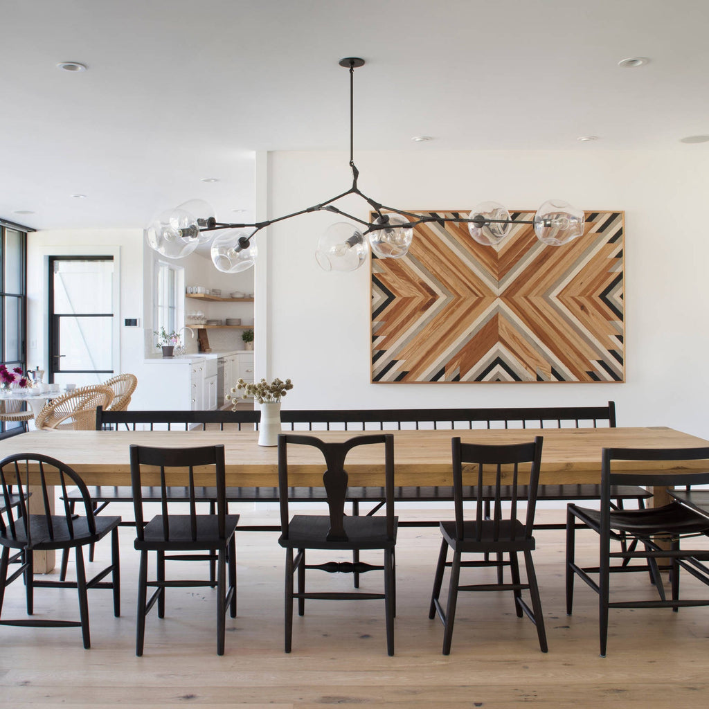 A black-armed Clérachic chandelier extends over a long wooden dining table, with a wooden geometric panel on the wall behind it.